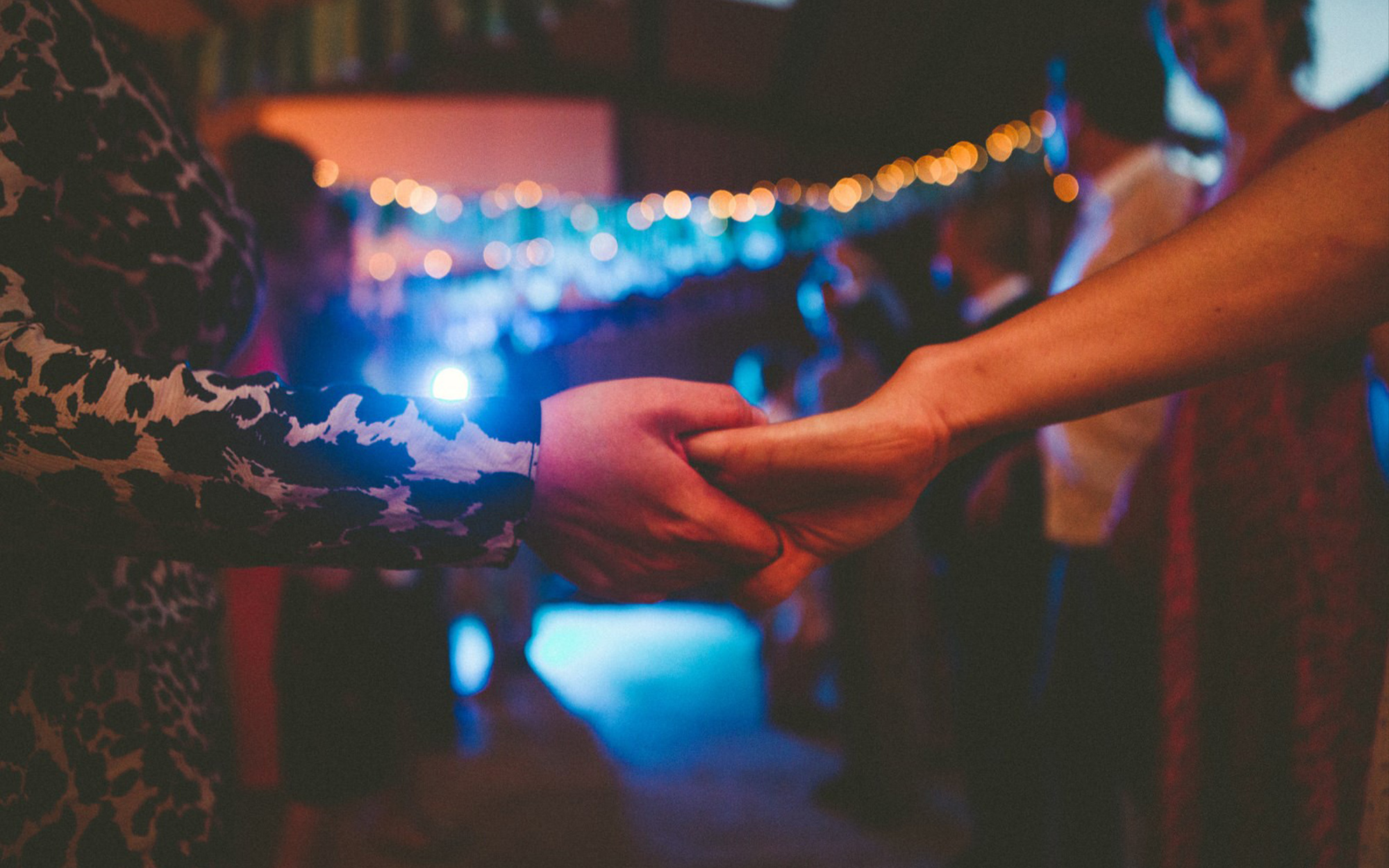 A close up image of two people holding hands as if to dance. Behind them we can see a crowd of other people and distant lights.